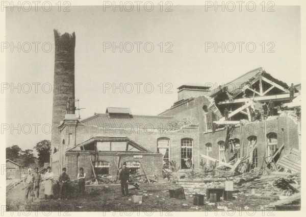Collapsed cotton mill in Nagoya, page, photomechanical print, Nagoya, cliché maker: Kazumasa Ogawa, Japan, in or after 1891-10-28 - in or before 1892, photographer, Chubu, paper, collotype, photoprint, height 202 mm x width 289 mm, collapsing building, consequences of an earthquake, Mino–Owari earthquake 1891, A large brick building with a tall cylindrical chimney stands against a pale sky, part of the structure showing a collapsed roof and exposed wooden beams and rafters; broken brickwork, scattered timber, and debris cover the ground in front of the building, where several people stand among the rubble, some on the ruined upper floor and others at ground level; windows and arched openings in the brick facade are visible, along with a small pitched roof section and a rectangular rooftop vent; a few wooden crates and loose materials lie on the grassy area in the foreground.