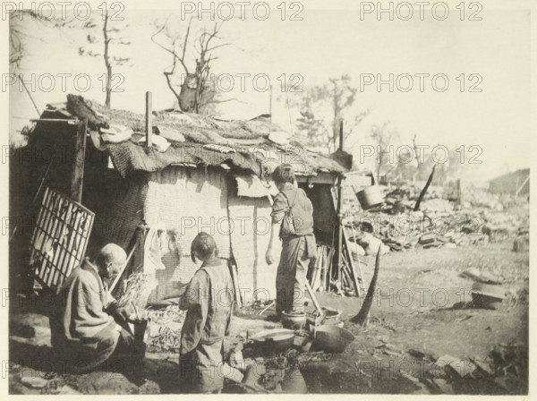 Two women and a man in front of a temporary home in Japan, page, photomechanical print, Japan, cliché maker: Kazumasa Ogawa, after 1891-10-28 - in or before 1892, photographer, paper, collotype, photoprint, height 213 mm x width 290 mm, housing, consequences of an earthquake, Mino–Owari earthquake 1891, A sepia-toned outdoor scene shows a makeshift shelter with a woven fabric covering and rough materials on its roof, supported by vertical poles and surrounded by scattered tools and containers. Several people stand or sit near the shelter, wearing simple clothing and facing the shelter or each other. Pots, pans, buckets, and other domestic items are arranged on the ground in front of the shelter. Leafless trees rise behind the shelter and a landscape of rubble and uneven ground extends into the background.