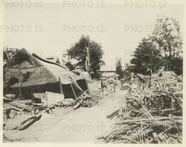 Destroyed buildings along the road to Gifu, page, photomechanical print, Japan, cliché maker: Kazumasa Ogawa, after 1891-10-28 - in or before 1892, photographer, paper, collotype, photoprint, height 218 mm x width 280 mm, public road, ruin of a building, architecture, consequences of an earthquake, Mino–Owari earthquake 1891, A wide dirt path runs through a scene of wooden debris and thatched roofs, with piles of planks and broken timber scattered along both sides; on the left a large thatched structure with irregular roof edges and nearby smaller wooden crates and frameworks sit amid rubble, while on the right stacks of wooden beams and lattice-like pieces form a cluttered foreground leading toward rudimentary wooden scaffolding and frames; in the middle distance a pair of cartlike objects with rounded fronts stand on the path flanked by more damaged wooden materials, and behind them a cluster of trees with leafy canopies rises above small rooflines and simple structures.