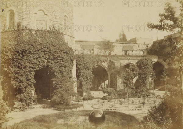 View of the cloister of the ruins of St. Bavo's Abbey in Ghent, Bruges, photograph, page, inscription: 'Gand ruines de l'abbaye de St Bavon', Ghent, c. 1862 - c. 1872, photographer, paper, albumen print, albumendruk, height 143 mm x width 201 mm, Photo on page 37., ruin of church, monastery, cloisters, Sint-Baafsabdij, A sepia-toned outdoor scene showing an old stone building with arched openings and climbing plants covering large portions of its walls and columns, lattice windows set into the stone, a courtyard with planted beds and low hedges, a small tree and scattered shrubs, a reflective spherical object on a stand positioned on a grassy area in the foreground, and a person partially visible standing near one of the vine-covered openings.