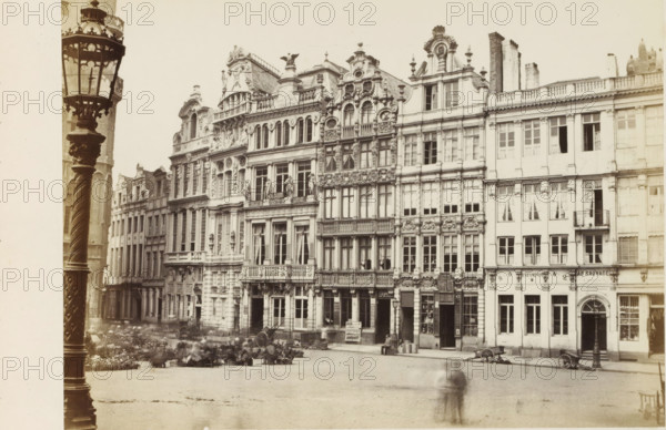 Buildings on the Grand Place in Brussels, Bruxelles - Maisons de la Grand'Place, photograph, page, name: '(Phot. de Brand frères, rue de la Madeleine, 32.)', photographer: Brand Frères, Grand Place (Brussels), c. 1862 - c. 1872, Brand Frères, photographer, Brussels, paper, albumen print, height 150 mm x width 204 mm, Photo on page 11., square, circus, façade (of house or building), A row of ornate multi-story buildings with decorative facades, sculpted figures, balconies, and many tall windows fills the background, with a tall ornate lamp post in the foreground at left. The buildings show varied architectural details including carved reliefs, rounded and arched window frames, and rooftop ornamentation. The street in front has scattered carts and barrels, blurred figures suggesting movement, and a small raised flowerbed or planter area near the center. The overall scene appears sepia-toned with high contrast between the pale sky and the detailed building facades.