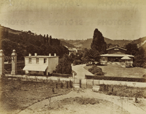 Chaudfontaine, Ladies' shoes and a view of Chaudfontaine, photograph, page, Above ladies' shoes and stockings, below a view of Chaudfontaine., Belgium, c. 1862 - c. 1872, photographer, paper, albumen print, height 403 mm x width 294 mm, Photos on page 27., shoes, sandals, city-view in general, 'veduta', stockings, socks, A sepia-toned view of a landscaped property with a gated entrance in the foreground and a gravel path leading inward past trimmed hedges and small trees toward buildings set among taller trees and rolling hills; on the left a two-story light-colored building with an awning and outdoor chairs sits beside a decorative columned gateway, while on the right a larger pavilion-like structure with a covered porch and a freestanding gazebo occupies a lawn area with scattered tables and chairs, and a tall conical tree rises behind the pavilion against a backdrop of wooded slopes and distant ridgelines under a pale sky.