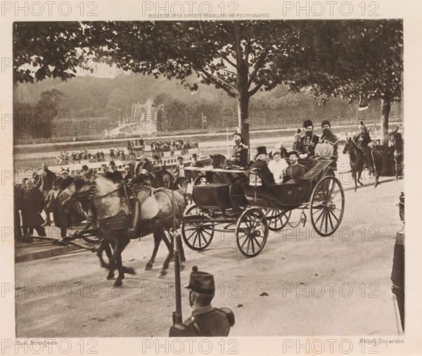 Carriage with Tsar Nicholas II of Russia and Tsarina Alexandra Feodorovna near Versailles, Sur la route de Versailles, page, photomechanical print, photographer: Bourgeois, France, cliché maker: Dujardin, c. 1896, clichémaker, paper, photoaquatint, height 131 mm x width 162 mm, Person, Nicholas II Alexandrovich (Tsar of Russia), Alexandra Feodorovna (Tsarina of Russia), ruler, sovereign, sovereign - BB - female ruler, passenger carriage, A sepia-toned scene shows a street with horse-drawn carriages and mounted riders beneath large leafy trees, with a carriage in the foreground carrying several formally dressed passengers including a woman in a light dress and men in dark attire, drivers and uniformed attendants nearby, horses in harness moving along a paved road, groups of onlookers and additional horsemen in the background, and a distant landscape of trees and a structure partially visible beyond a body of water.