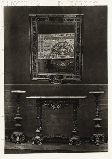 Table, two candle holders and a mirror, page, photomechanical print, name: 'De Goupil & Cie Imp. Photoglytique Paris', number: '477 478 479', photographer: Goupil & Cie., Paris, 1870, paper, Woodburytype, Woodburytypie, height 191 mm x width 134 mm, candlestick, mirror, A framed rectangular mirror with an ornate patterned border hangs on a plain dark wall, reflecting a blurred decorative textile and part of an ornamental object; below the mirror a narrow table with a carved apron and turned legs stands centered between two tall ornate candlesticks with wide circular tops and elaborately carved pedestal bases, all resting on a flat surface.