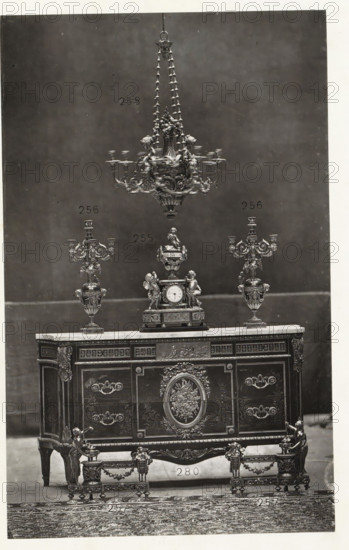 Chest of drawers with a clock and two candlesticks in Louis XVI style, page, photomechanical print, name: 'De Goupil & Cie Imp. Photoglytique Paris', number: '258 256 255 280 257', photographer: Goupil & Cie., Paris, 1870, paper, Woodburytype, Woodburytypie, height 195 mm x width 122 mm, candlestick, clock, ornamental cupboard, A tall ornate chandelier hangs from the top center, featuring multiple tiers, decorative chains, and sculpted floral and scroll details, positioned above a matching decorative clock and flanked by two elaborate candelabra with curved arms and ornamental bases. The clock sits on a carved rectangular pedestal and includes sculpted figures and ornamental vases as part of its design. All objects rest on a richly decorated wooden chest of drawers with a light-colored top, intricate paneling, carved floral medallions, decorative handles, and gilt-like embellishments on the corners and legs. In front of the chest are matching low stands with ornate legs, carved garland...