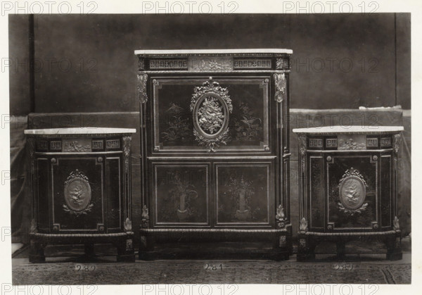 Secretary and two triangular corner pieces of furniture decorated with medallions and plant motifs, page, photomechanical print, name: 'De Goupil & Cie Imp. Photoglytique Paris', number: '281 282', photographer: Goupil & Cie., Paris, 1870, paper, Woodburytype, Woodburytypepie, height 123 mm x width 183 mm, writing-table, writing-desk, ornamental cupboard, A central ornate cabinet with a flat top sits between two smaller matching cabinets, all featuring dark wood panels with lighter tops and decorative molding; the central cabinet has an oval floral relief surrounded by carved garlands on its upper door and framed panels with painted floral motifs on its lower doors, while each side cabinet displays a similar oval relief on its front panel and decorative corner carvings, all standing on short carved legs against a plain dark backdrop with a lighter floor beneath.