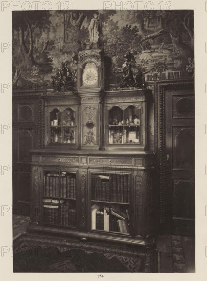 Room in the house of Antonin Terme with oak cupboard, photograph, page, Liège, c. 1880 - in or before 1885, photographer, paper, platinum process, height 223 mm x width 154 mm, art collection, art gallery, cabinet of curiosities, 'Kunst- und Wunderkammer', An ornate wooden cabinet with carved panels and glass-fronted upper compartments fills the image, the upper central section topped by a decorative clock face and a small statue, flanked by sculpted horse figures; the glass compartments display various small objects and vessels, while the lower sections reveal shelves of books and stacked papers behind glass doors; the cabinet is set against a wall covered with a detailed tapestry or mural depicting trees, foliage, and a swan in water, and parts of carved wooden doors with decorative moldings are visible at the sides, a patterned rug lies on the floor in front.