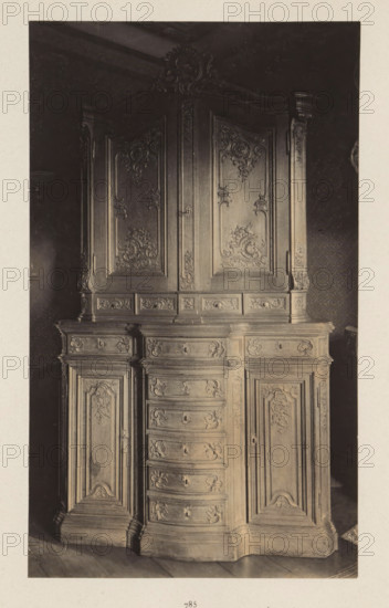 Decorated cupboard from the collection of Antonin Terme, photograph, page, Liège, c. 1880 - in or before 1885, photographer, paper, platinum process, height 224 mm x width 154 mm, art collection, art gallery, cabinet of curiosities, 'Kunst- und Wunderkammer', A tall wooden cabinet with ornate carved panels fills the frame, showing an upper section with two large doors adorned with intricate floral and scrollwork carvings and a decorative carved crest at the top, and a lower section with a central column of curved drawers flanked by paneled cupboard doors, all surfaces featuring detailed relief ornamentation and molded edges, set against a dark patterned wall and resting on a wooden floor.