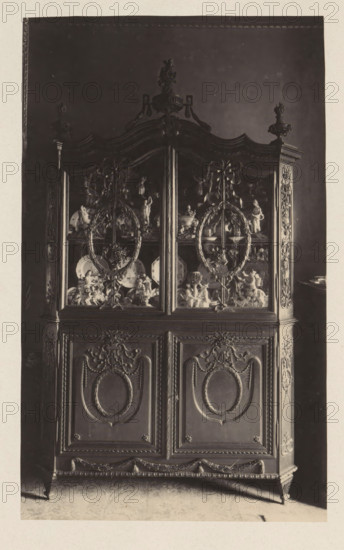 Cupboard in the house of Antonin Terme with statues and plates, photograph, page, Liège, c. 1880 - in or before 1885, photographer, paper, platinum process, height 210 mm x width 154 mm, art collection, art gallery, cabinet of curiosities, 'Kunst- und Wunderkammer', A tall ornate wooden cabinet with carved decorative panels and mouldings fills the frame, featuring an upper section with glass doors revealing shelves of ceramic and sculptural objects, including plates and figurines, and circular wreath-like carved motifs on the glass; the top of the cabinet has a curved, scalloped crest with a central sculpted finial and smaller carved finials at the corners, while the lower section shows recessed panels with oval medallion carvings, ribbon and floral ornamentation, and a carved apron and short tapered legs resting on a patterned floor.