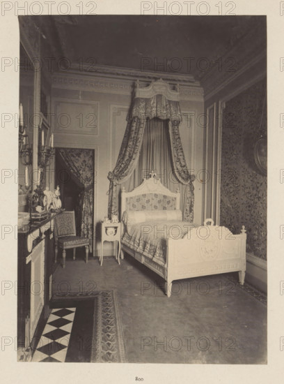 Room in the house of Antonin Terme with decorated bed, photograph, page, Liège, c. 1880 - in or before 1885, photographer, paper, platinum process, height 223 mm x width 161 mm, bed, art collection, art gallery, A photograph of an ornate bedroom showing a carved wooden bed with a decorative headboard and footboard, dressed with patterned bedding and a cylindrical bolster, positioned beneath a fabric canopy with heavy draped curtains tied back at the sides; beside the bed is a small painted bedside table holding decorative items and a chair with upholstered seat and carved legs sits near a mantelpiece topped with candlesticks and ornamental figurines; walls feature molded panels and patterned wallpaper, an ornate ceiling cornice runs along the top, and a patterned rug and a tiled border cover the floor.
