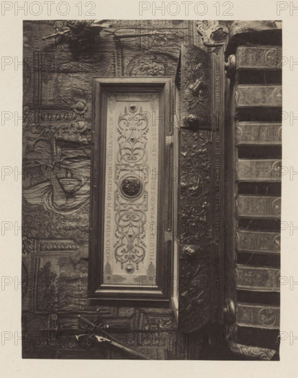 Virginal from the collection of Antonin Terme, photograph, page, Liège, c. 1880 - in or before 1885, photographer, paper, platinum process, height 149 mm x width 196 mm, musical instruments, group of musical instruments, art collection, art gallery, A vertical photograph shows a framed rectangular panel with ornate carved designs and a circular medallion at its center, mounted against a richly textured tapestry or wall covering. To the right of the frame is a carved wooden structure with elaborate reliefs of scrolling foliage and figures, and adjacent to that are stacked fabric-covered books or cushions with patterned bands. To the left of the framed panel a draped figure is partly visible within the tapestry, and the lower area shows decorative architectural details and a long narrow object resembling a pole or staff resting diagonally. The image is rendered in sepia tones with high contrast highlighting the carved and textile textures.