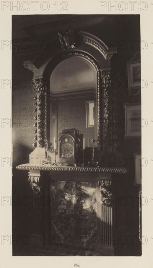 Chimney with clock in the house of Antonin Terme, photograph, page, Liège, c. 1880 - in or before 1885, photographer, paper, platinum process, height 217 mm x width 116 mm, clock, art collection, art gallery, chimney-piece, mantel piece, A tall ornate mirror with a carved arch and decorative molding sits above a mantelpiece, the mirror reflecting part of a patterned wall and a framed picture. On the mantel rests an elaborate clock with a domed top and a round face flanked by a pair of candlesticks, and the mantel edge features a carved rope-like trim. The fireplace surround displays intricate carved columns and floral or foliate designs on the panel below the mantel, and the surrounding wall shows patterned wallpaper and framed artwork. The overall tones are dark and the image appears monochrome.