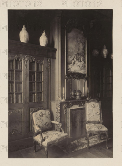 Room with chimney and two bookcases in the house of Antonin Terme, photograph, page, Liège, c. 1880 - in or before 1885, photographer, paper, platinum process, height 208 mm x width 147 mm, art collection, art gallery, chimney-piece, mantelpiece, bookcase, with doors, A sepia photograph showing an ornate interior corner with carved wooden cabinetry and a tall bookcase with glass-paneled doors filled with books, fluted decorative columns, and sculpted floral motifs; two large lidded ceramic urns sit on top of the cabinetry; a carved mantel with decorative vases and tall candlesticks stands beneath a framed painting depicting a pastoral scene; two upholstered chairs with floral fabric and carved wooden frames are positioned near the mantel on a patterned rug; additional decorative ceramic vessels are visible on a high shelf in the darker background.
