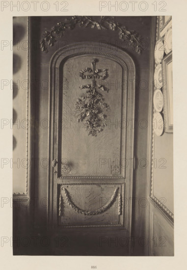 Wooden decorated door in the house of Antonin Terme, photograph, page, Liège, c. 1880 - in or before 1885, photographer, paper, platinum process, height 222 mm x width 150 mm, door, art collection, art gallery, sculpture (relief, sculpture), A tall wooden door with an arched top occupies the center of the image, featuring carved decorative elements including a ribbon-tied floral swag near the top and a carved garland draped across the lower panel, framed by beaded molding and corner rosettes; to the right of the door a narrow wall section displays a vertical row of decorative plates mounted beside a framed picture, and to the left a portion of an adjacent wall and molding is visible, all shown in a monochrome tone.