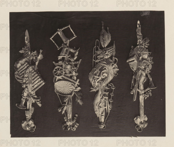 Four small trophies of music, fish and agriculture in giltwood from the collection of Antonin Terme, photograph, page, Liège, c. 1880 - in or before 1885, photographer, paper, platinum process, height 155 mm x width 191 mm, trophy, ie: decorative arrangement of symbolic or typical objects, ornament, art collection, art gallery, A rectangular dark panel centered on a lighter background holds four vertical ornamental tassel-like sculptures, each composed of carved objects and natural motifs; the leftmost piece includes a trumpet, a bundle of rods, leaves, and ribbon tied at the bottom, the second from left features a woven basket, a small wheel, floral sprigs, a broken chair frame, and dangling foliage, the third shows a carved fish, coils of rope, woven textures, and a twisted tassel ending in a rosette, and the rightmost assembly contains a horn, a shield-like form, and a blade-like element rising upward, all rendered in a monochrome, high-contrast photographic style against a subtly g...