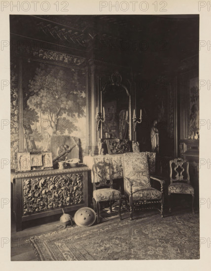Room in the house of Antonin Terme with his collection of antiquities, photograph, page, Liège, c. 1880 - in or before 1885, photographer, paper, platinum process, height 216 mm x width 167 mm, interior of the house, art collection, art gallery, A richly furnished interior with ornate carved wooden furniture and patterned upholstery, including a pair of high-backed chairs and a smaller chair arranged near a table covered with a decorative cloth; a large carved cabinet topped with small framed panels and objects sits beneath a tapestry depicting trees and foliage that covers a wall; a tall mirror with an elaborate frame and flanking candle sconces stands behind the table, reflecting parts of the room; a sculpted figurine or draped statue is visible in a darker corner; decorative shields and a helmet rest on the floor near the carved cabinet, and a patterned rug covers the floor in the foreground.