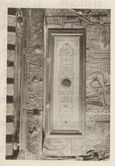 Virginal Duke of Cleves from the collection of Antonin Terme, page, photomechanical print, annotation: 'Virginale - ouvert vue ... collection A. Terme, 128 =', c. 1880 - in or before 1885, paper, collotype, photoprint, height 137 mm x width 203 mm, Print is loose in the book., spinet, virginal, art collection, art gallery, A grayscale photograph of an ornate carved doorway or panel rotated sideways, featuring an elaborate rectangular central panel with symmetrical scrollwork, circular medallions, and decorative relief elements, flanked by richly carved vertical borders with sculpted figures, faces, and heraldic motifs; to the right of the central panel part of a draped figure holding fabric is visible against a patterned wall, and above and below are architectural details including columns, sculpted friezes, and decorative moldings, with a narrow margin of blank pale paper around the image.