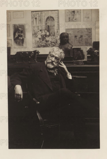 Portrait of Antonin Terme, photograph, page, c. 1880 - in or before 1885, photographer, paper, platinum process, height 186 mm x width 122 mm, Person, Antonin Terme, historical persons, An older man with a beard and mustache sits in a dark suit and patterned neckerchief, leaning his head on one hand and resting the other arm on the chair, with a contemplative expression; behind him a wooden surface holds several sculptures including a dark bust of a girl and a dark animal figure, and the wall behind displays framed artworks and illustrations in various sizes.