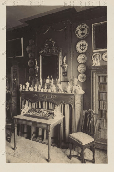 Room in the house of Antonin Terme with his collection of antiquities, plates and statues, photograph, page, Liège, c. 1880 - in or before 1885, photographer, paper, platinum process, height 227 mm x width 147 mm, interior of the house, dish, plate, saucer, art collection, art gallery, A sepia-toned interior scene showing an ornate mantelpiece topped with numerous small figurines, a clock, and decorative vases, with a large framed mirror above it flanked by mounted round decorative plates on the wall and small statue wall brackets; in front of the mantel is a rectangular table with a tray of objects and two small sculpted groups at its corners, a carved wooden chair with an upholstered seat positioned to the right, and a tall bookcase filled with volumes at the far right, while carved paneling, drapery beneath the mantel, patterned floor covering, and additional framed decorative elements are visible around the room.