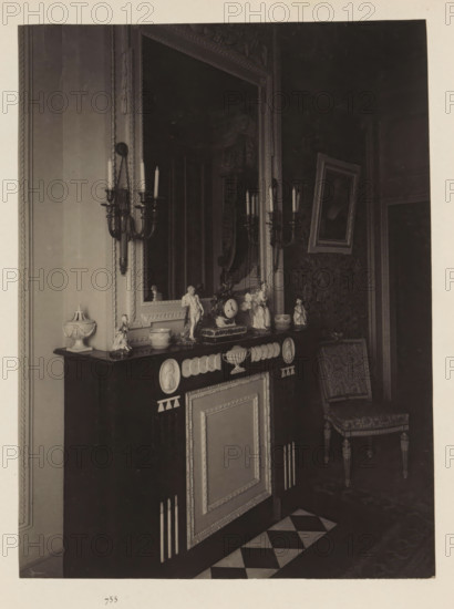 Room in the house of Antonin Terme with its collection of antiquities, statue of Hercules, clock and candlesticks, photograph, page, Liège, c. 1880 - in or before 1885, photographer, paper, platinum process, height 220 mm x width 165 mm, interior of the house, art collection, art gallery, A dark mantel with decorative panels and medallion reliefs holds various small porcelain and sculpted figurines and a central clock, flanked by two ornate wall sconces with candles; above the mantel is a large framed mirror with an embellished frame reflecting part of the room, and to the right a framed picture is hung at an angle above a patterned chair with carved legs; the floor in front of the mantel shows a checkerboard tile strip and the surrounding walls feature moulding and patterned wallpaper.