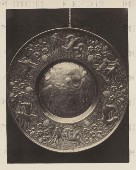 Copper bowl from the collection of Antonin Terme, photograph, page, Liège, c. 1880 - in or before 1885, photographer, paper, platinum process, height 205 mm x width 163 mm, dish, plate, saucer, art collection, art gallery, birth and youth of Christ, A round metallic plate with a raised rim hangs by a short chain against a dark rectangular background, the rim and outer band decorated with intricate reliefs of floral clusters, rounded textured fruits, and various stylized animals and humanlike figures in dynamic poses; the central circular panel contains a finely detailed scene showing several humanlike figures gathered around a seated figure holding a small child, with other standing and kneeling figures nearby and architectural and decorative motifs in the background.