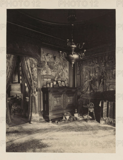 Room in the house of Antonin Terme with his collection of antiquities, wooden spinning wheel and a cupboard, photograph, page, Liège, c. 1880 - in or before 1885, photographer, paper, platinum process, height 215 mm x width 165 mm, 'Kunstschrank', art collection, art gallery, other rooms, An ornate interior scene showing a richly decorated room with carved wooden furniture, patterned tapestries covering the walls, and heavy draped curtains; a large metallic chandelier with curved arms hangs from the ceiling above a carved cabinet topped with small decorative objects and framed items; to the right a tapestry panel depicts robed figures and architectural motifs, and a small spinning wheel or similar wooden apparatus stands on the floor nearby; the floor is covered by a worn patterned rug, and various fabrics, cushions, and a small chair with carved legs are visible near an open doorway leading to another curtained space.