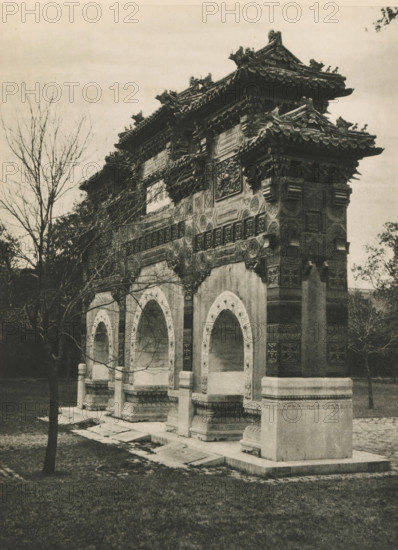 Paifang at the Confucius Temple in Beijing, page, photomechanical print, photographer: Donald Mennie, Beijing, cliché maker: anonymous, c. 1915 - in or before 1920, paper, rotogravure, height 275 mm x width 203 mm, gate, entrance, Confucius Temple, A stone archway structure with ornate carved details and a tiled roof featuring decorative ridge figures stands in a grassy area, framed by bare-branched trees; the structure has multiple arched openings set on raised bases with carved supports and patterned panels, and steps and stone slabs lead up to the openings, while the weathered surface and intricate relief work are visible under an overcast sky.