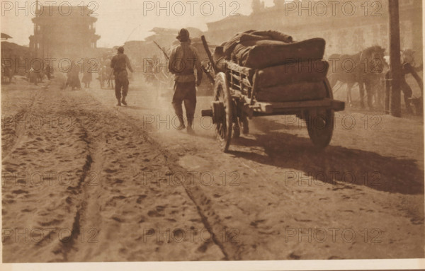 Away with horse-drawn carts, page, photomechanical print, photographer: Donald Mennie, Beijing, cliché maker: anonymous, c. 1915 - in or before 1920, paper, rotogravure, height 183 mm x width 278 mm, two-wheeled, animal-drawn vehicle, A dusty street scene in sepia tones showing a dirt road with deep ruts and footprints in the foreground, a wooden cart loaded with large bundled sacks seen from behind and slightly to the side, the cart’s wheel and axle visible and casting a shadow, two men walking ahead of the cart wearing loose clothing and hats, several animals harnessed at the side near a post, indistinct groups of people and figures further down the street, and a large gate-like structure silhouetted against a bright sky in the background.
