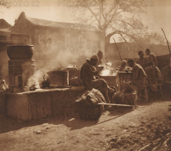 Unknown eating men, page, photomechanical print, photographer: Donald Mennie, Beijing, cliché maker: anonymous, c. 1915 - in or before 1920, paper, rotogravure, height 217 mm x width 239 mm, historical persons, eating, A sepia-toned outdoor scene showing a group of people gathered around steaming pots and cooking fires near a low stone wall, with large metal cauldrons and barrels placed on and near the wall; some figures are seated while others stand or lean, several are engaged with the steaming pots, and a person is carrying a load on a pole; in the background there is a building with a tiled roof and decorative gables, a tall tree with branches spreading overhead, and a curved archway set into a wall, all seen through hazy smoke.