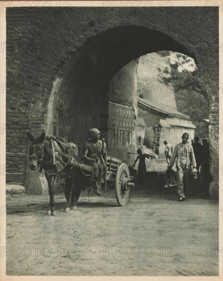 Unknown persons with horse and cart at a gate, page, photomechanical print, photographer: Donald Mennie, Beijing, cliché maker: anonymous, c. 1915 - in or before 1920, paper, rotogravure, height 252 mm x width 203 mm, historical persons, two-wheeled, animal-drawn vehicle, gate, entrance, A wide stone archway framed by textured brickwork opens onto a shadowed passage where a cart hitched to a donkey stands in front of the arch, the donkey facing forward with visible harnesses and ropes, its ears prominent; a partially shirtless figure sits on the cart holding a pole and wearing a cloth wrapped around the head; behind and within the arch a person balances a carrying pole across the shoulders with buckets hanging from its ends; another man walks forward carrying items in each hand and wearing light clothing; additional silhouetted figures stand further back in the passage; architectural details of worn walls, a wooden gate, and stone structures are visible through the arch, and a tree can...