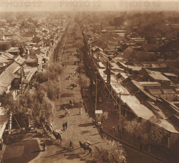 Street view in Beijing, page, photomechanical print, photographer: Donald Mennie, Beijing, cliché maker: anonymous, c. 1915 - in or before 1920, paper, rotogravure, height 218 mm x width 237 mm, street, A high vantage street view shows a long straight road flanked by rows of low buildings with sloping tiled roofs and awnings, trees lining parts of the street, overhead utility poles and wires along one side, and groups of people and animal-drawn carts moving along the roadway; the scene is captured in a sepia tone with distant rooftops and treetops fading into a hazy background.