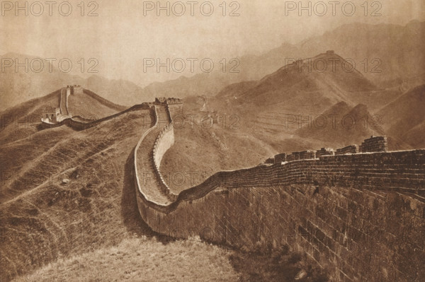 Great Wall of China, page, photomechanical print, photographer: Donald Mennie, Beijing, cliché maker: anonymous, c. 1915 - in or before 1920, paper, rotogravure, height 194 mm x width 280 mm, walls, fortified city, A long stone wall with battlements winds along a series of rolling, grassy hills receding into misty mountains, with several watchtowers and fortified structures punctuating the wall; the scene is rendered in a sepia tone, showing textured stonework on the wall faces, paths along the wall, undulating slopes, and distant layered ridgelines fading into the background.