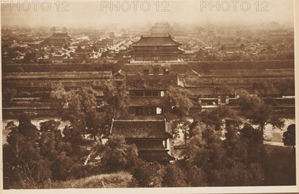 View of the Forbidden City in Beijing, page, photomechanical print, photographer: Donald Mennie, Beijing, cliché maker: anonymous, c. 1915 - in or before 1920, paper, rotogravure, height 182 mm x width 276 mm, city-view in general, 'veduta', Forbidden City, A sepia-toned aerial view shows a dense cluster of traditional tiled-roof buildings and pavilions arranged behind multiple long horizontal walls, with large roofed halls rising toward the background and rows of smaller peaked roofs filling the midground; trees and leafy vegetation appear in the foreground interspersed with pathways and courtyard areas, while the distant upper portion of the scene is hazy and shows further layered rooftops fading into the background.