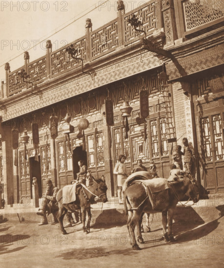Unknown persons and two donkeys in front of a shop in Beijing, page, photomechanical print, photographer: Donald Mennie, Beijing, cliché maker: anonymous, c. 1915 - in or before 1920, paper, rotogravure, height 240 mm x width 203 mm, historical persons, shop, store, ass, donkey, A sepia-toned street scene showing an ornate building facade with carved wooden panels, latticework railings along the upper level, decorative brackets and hanging lanterns, and a row of detailed doors and windows; several people stand and sit on the building's raised stone platform while others walk in front of it; two laden pack animals occupy the foreground with harnesses and cloth coverings; shadows fall across the ground and the lower part of the facade.