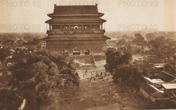 Gu Lou in Beijing, page, photomechanical print, photographer: Donald Mennie, Beijing, cliché maker: anonymous, c. 1915 - in or before 1920, paper, rotogravure, height 183 mm x width 279 mm, landscape with tower or castle, Gu Lou, A large multi-tiered traditional building with sweeping tiled roofs stands centrally, elevated on a wide base with arched openings at its ground level and an open gallery on the upper story. In front of the building a broad dirt pathway slopes downward through an open area where small groups of people are scattered, some standing and some walking. Trees with dense foliage frame the foreground and sides, and low rooftops and clustered buildings extend outward behind and beside the central structure. The image has a sepia tone and a hazy distant horizon with indistinct shapes of more trees and structures.