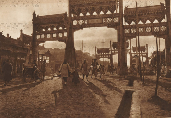 Paifang in Beijing, page, photomechanical print, photographer: Donald Mennie, Beijing, cliché maker: anonymous, c. 1915 - in or before 1920, paper, rotogravure, height 198 mm x width 278 mm, gate, entrance, A wide street scene in a sepia tone shows large ornate gate structures with carved lattice panels spanning the roadway, casting shadows on the uneven ground. People walk and stand beneath and around the gates, some wearing long coats and carrying items, others pulling or riding simple rickshaws. Shadows from the figures stretch along the cobblestone surface toward the foreground. Flanking buildings with pitched roofs and decorative eaves line the sides of the street, and utility poles rise between the gate openings. The overall lighting is low and diffuse, creating a muted, aged appearance.