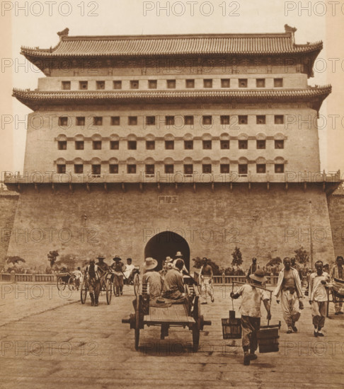 View of Zhengyang Men, page, photomechanical print, photographer: Donald Mennie, Beijing, cliché maker: anonymous, c. 1915 - in or before 1920, paper, rotogravure, height 245 mm x width 216 mm, gate, entrance, Zhengyang Men, A large multi-tiered stone and brick gatehouse with tiled overhanging roofs and rows of small square openings dominates the upper part of the image, while a wide arched passage sits at its center; in front of the gatehouse a paved plaza shows numerous people walking and working, some pulling or riding in two-wheeled rickshaws and carts, one cart with two seated figures viewed from behind, others carrying buckets suspended from a shoulder pole, and a few standing or walking individuals wearing hats and loose clothing.