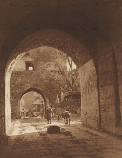 View of two gates in Beijing, page, photomechanical print, photographer: Donald Mennie, Beijing, cliché maker: anonymous, c. 1915 - in or before 1920, paper, rotogravure, height 257 mm x width 203 mm, gate, entrance, A sepia-toned view through a series of brick archways opening onto a street scene, with the nearest archway framing a passageway floor showing wet patches and a stone in the foreground. Beyond the arch, a person walks away leading a donkey on a rope, other figures and animals are visible further down the street, and a small wooden shelter or stall stands to one side. Trees with bare branches rise above the far wall, and the brickwork of the arches and adjoining walls shows texture and wear. Light filters through the arches creating areas of shadow and brightness along the ground and walls.