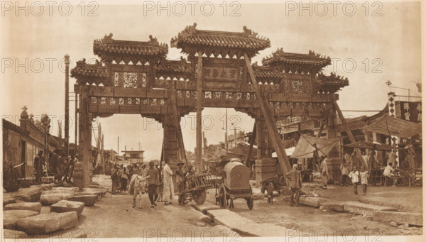 Street view with paifang, page, photomechanical print, photographer: Donald Mennie, Beijing, cliché maker: anonymous, c. 1915 - in or before 1920, paper, rotogravure, height 167 mm x width 289 mm, street, gate, entrance, A wide street scene dominated by a large ornate wooden gateway with multiple tiled roofs and decorative carvings, supported by tall pillars and angled braces; people move beneath and around the gateway, including individuals walking, standing, and conversing, some leading or standing by animals and simple carts; market stalls and temporary canopies line the street to the right with groups of people near them, while low stone slabs and building facades border the left side; the overall image has a sepia tone and shows a busy urban environment with architectural details and everyday street activity.