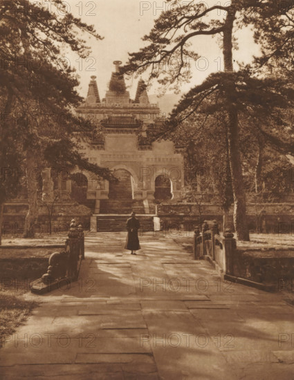 Unknown man at a temple, page, photomechanical print, photographer: Donald Mennie, Beijing, cliché maker: anonymous, c. 1915 - in or before 1920, paper, rotogravure, height 259 mm x width 204 mm, historical persons, adult man, temple, shrine, non-Christian religions, Temple of Azure Clouds, A sepia-toned photograph showing a stone pathway leading toward a large ornate building framed by tall trees, with a person wearing a long dark coat standing on the path facing the building; the building features arched doorways, tiered roof details, and stairways, while stone balustrades and carved posts line the sides of the path and trees with spreading branches partially obscure the view.