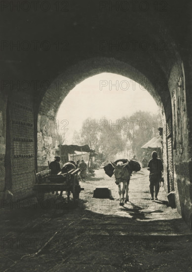 Gate in Beijing, page, photomechanical print, photographer: Donald Mennie, Beijing, cliché maker: anonymous, c. 1915 - in or before 1920, paper, rotogravure, height 279 mm x width 200 mm, historical persons, ass, donkey, gate, entrance, A dark stone archway frames a sunlit scene beyond, with the arch's textured interior visible on both sides; under the arch a narrow dirt path leads outward where a pack animal with saddlebags walks toward the viewer accompanied by a person walking beside it; to the left a wooden cart and another person are partly visible in shadow, and to the right the arch wall shows rough brick and a narrow doorway; beyond the arch silhouettes of trees rise in bright haze and several small canopies or stalls and faint figures are visible in the light.