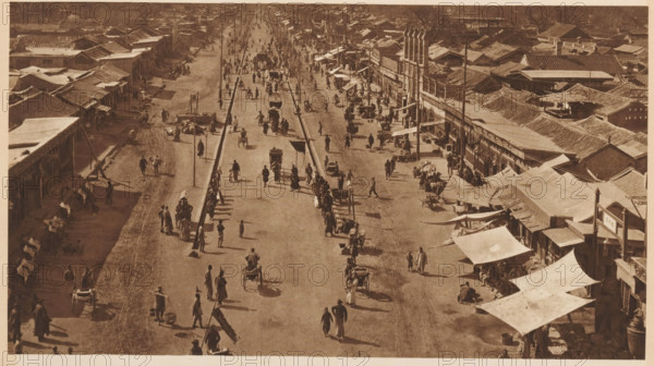 Street view in Beijing, page, photomechanical print, photographer: Donald Mennie, Beijing, cliché maker: anonymous, c. 1915 - in or before 1920, paper, rotogravure, height 164 mm x width 290 mm, street, A sepia-toned aerial view of a wide street lined with low buildings and awnings on both sides, with a raised central divider and lamp posts running down its length, showing many people walking or standing, several handcarts and rickshaws moving along the roadway, and groups of figures gathered near the storefronts and under fabric canopies, with tiled roofs extending into the distance and long shadows cast across the ground.