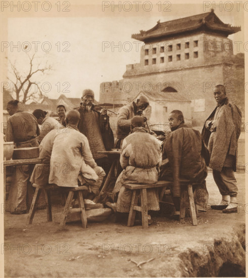 Group of unknown men, some of whom smoke pipes, page, photomechanical print, photographer: Donald Mennie, Beijing, cliché maker: anonymous, c. 1915 - in or before 1920, paper, rotogravure, height 242 mm x width 217 mm, historical persons, adult man, pipe, tobacco, A sepia-toned scene shows a group of people gathered around a low wooden table with simple stools, most seated with their backs or sides to the camera and wearing layered, textured clothing and wrapped garments. Two older men stand near the table, one playing a long pipe and another leaning over a large pot or basin while holding a utensil. A tall man stands to the right wearing a long quilted coat and looking toward the camera. In the background a multi-tiered brick building with a tiled roof and rows of small square openings rises above the group, and bare tree branches frame the left side of the image. The ground in the foreground is bare earth with scattered small debris and rough stone edging visible at the bottom right.