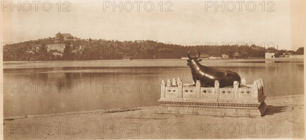 View of the Winter Palace in Beijing, page, photomechanical print, photographer: Donald Mennie, Beijing, cliché maker: anonymous, c. 1915 - in or before 1920, paper, rotogravure, height 178 mm x width 278 mm, trees, palace, A sepia-toned scene of a large body of water with a shoreline in the foreground and a hillside with buildings and trees across the water, with a dark, polished statue of a reclining bull placed on an ornate rectangular stone platform near the water’s edge; the platform has carved railings and decorative details, and the bull faces toward the water with curved horns and a smooth surface, while the distant shore shows pagoda-like structures and clustered foliage under a pale sky.