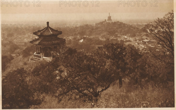 Bronze sculpture of an ox on a river near the Summer Palace in Beijing, page, photomechanical print, photographer: Donald Mennie, Beijing, cliché maker: anonymous, c. 1915 - in or before 1920, paper, rotogravure, height 138 mm x width 289 mm, sculpture, river, Summer Palace, A sepia-toned elevated view showing a traditional multi-tiered pavilion with a curved tiled roof perched on a hillside, surrounded by dense trees and foliage in the foreground, with a distant landscape of low buildings and another large stupa-like structure on a hill shrouded in haze, and a bare-branched tree framing the right edge of the scene.