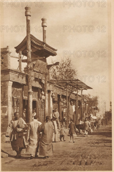 Street view in Beijing, page, photomechanical print, photographer: Donald Mennie, Beijing, cliché maker: anonymous, c. 1915 - in or before 1920, paper, rotogravure, height 279 mm x width 188 mm, street, A sepia-toned street scene shows a row of low buildings with ornate eaves and wooden columns supporting a projecting roof structure; tall carved posts rise above the roofline topped with capped finials. People in traditional loose garments walk and stand along the unpaved road, including adults and children, some carrying bundles or holding small objects. Market-style awnings and simple wooden frameworks extend from the building fronts, and hanging items are visible near doorways. A few animals are present among the people toward the middle distance. Trees rise behind the buildings and the sky appears open and mostly clear above the scene.