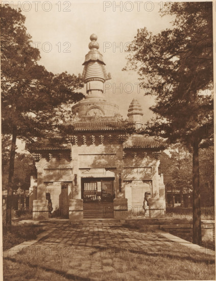 Stupas at a temple in Beijing, page, photomechanical print, photographer: Donald Mennie, Beijing, cliché maker: anonymous, c. 1915 - in or before 1920, paper, rotogravure, height 263 mm x width 203 mm, places and objects of worship, non-Christian religions, A sepia-toned photograph of a stone gateway topped by a multi-tiered ornamental tower framed by tall trees on both sides, with a paved path and steps leading up to the entrance flanked by carved stone pillars and guardian statues, decorative eaves and carved reliefs visible on the gateway structure, and shaded foliage creating dappled light across the foreground.