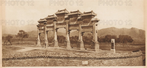 Paifang at the imperial tombs of the Ming dynasty in Beijing, page, photomechanical print, photographer: Donald Mennie, Beijing, cliché maker: anonymous, c. 1915 - in or before 1920, paper, rotogravure, height 139 mm x width 288 mm, gate, entrance, A wide sepia-toned photograph shows a large ornate stone archway with multiple vertical pillars supporting horizontal beams and decorative tiled roofs, each roof section featuring detailed eaves and sculpted ridge ornaments; the bases of the pillars rest on carved stone pedestals with relief designs. The archway stands on a raised stone platform behind a low brick wall with a patterned band near its top. In front of the archway on the platform a person is visible carrying a yoke with hanging baskets. Leafless trees and taller trees occupy the landscape behind and around the structure, and low rolling hills and a distant jagged mountain ridge form the background under a pale sky. A freestanding carved stone marker stands near the right side o...