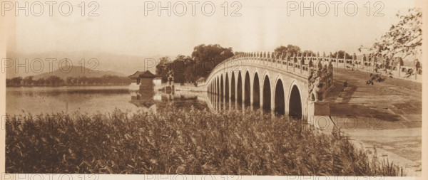 Bridge at the Summer Palace in Beijing, page, photomechanical print, photographer: Donald Mennie, Beijing, cliché maker: anonymous, c. 1915 - in or before 1920, paper, rotogravure, height 124 mm x width 289 mm, river, bridge, A sepia-toned scene shows a curved stone bridge with multiple archways reflected in calm water, ornamental balustrades lining its sides, and sculpted figures at the near end of the bridge. Tall grasses fill the foreground along the water’s edge. On the far side of the bridge a pavilion with a pitched roof and adjacent small structures sit beside trees that extend along the shoreline. Hills rise in the distant background beneath a pale sky.