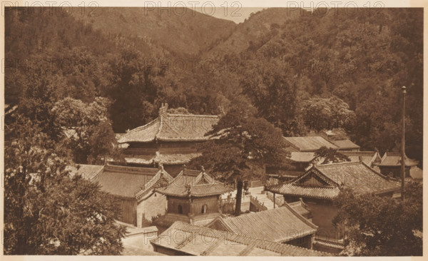 View of a landscape with mountains and roofs, page, photomechanical print, photographer: Donald Mennie, Beijing, cliché maker: anonymous, c. 1915 - in or before 1920, paper, rotogravure, height 172 mm x width 277 mm, roof (of house or building), mountains, A sepia-toned aerial view showing traditional tiled roofs and ornate eaves of a clustered complex of buildings surrounded by dense trees and hills, with layered rooftops, decorative ridgelines, a few tall trees rising among the structures, shadowed courtyards and pathways between the buildings, and a forested hillside forming the background.